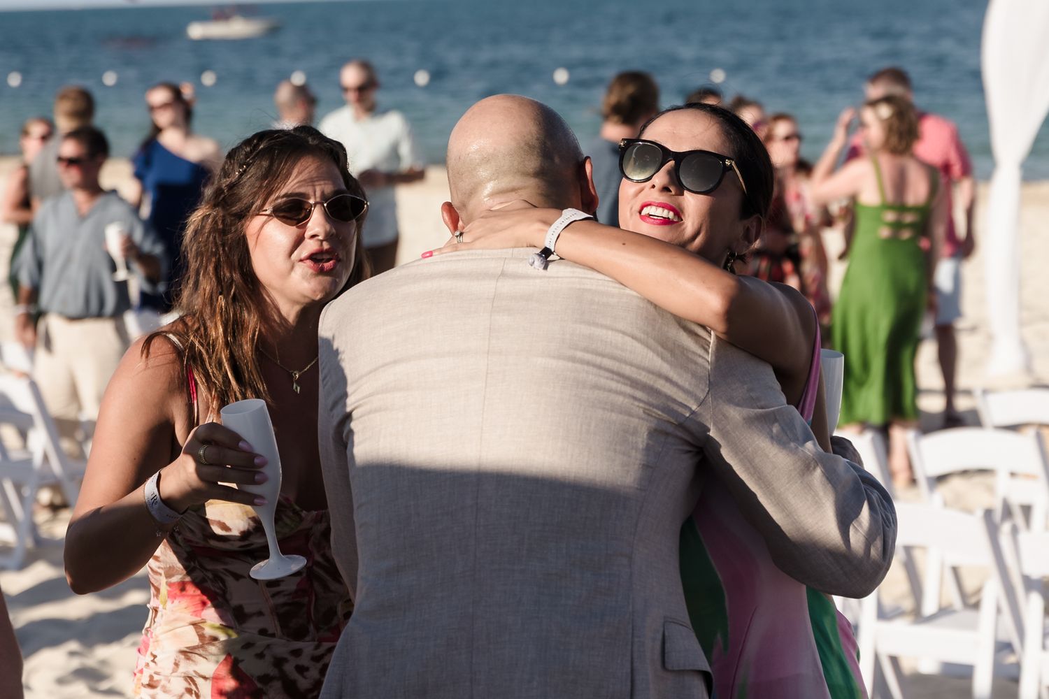 Guests embracing and celebrating at a sunny beach wedding ceremony with white chairs and ocean backdrop.