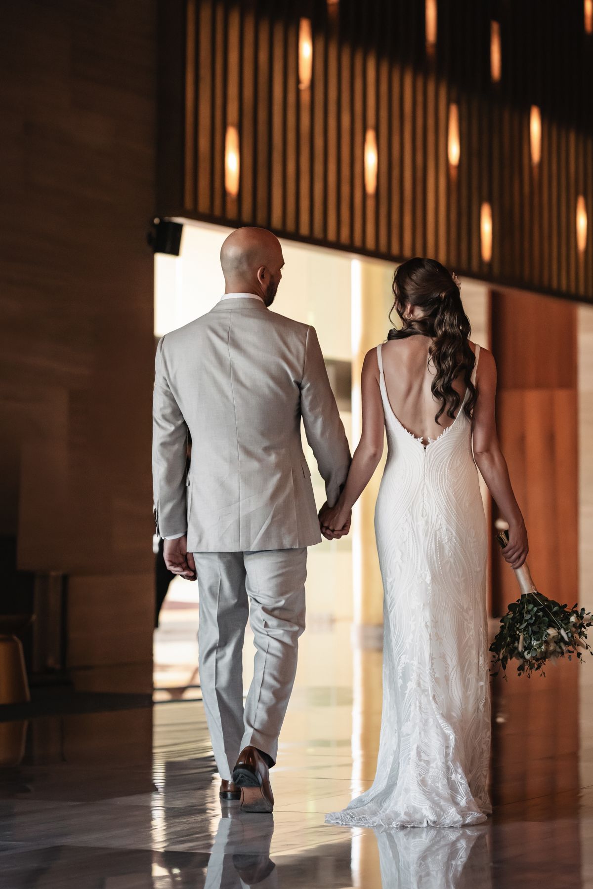 Bride and groom walking hand in hand, bride in backless lace gown holding bouquet, groom in light gray suit.