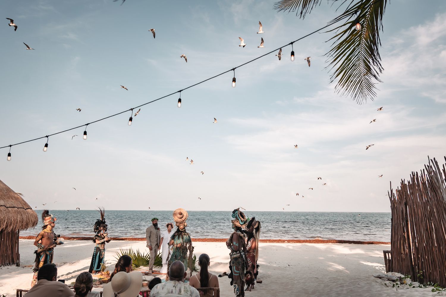 Beach ceremony with costumed performers, string lights, and seagulls flying over the ocean at a tropical venue.