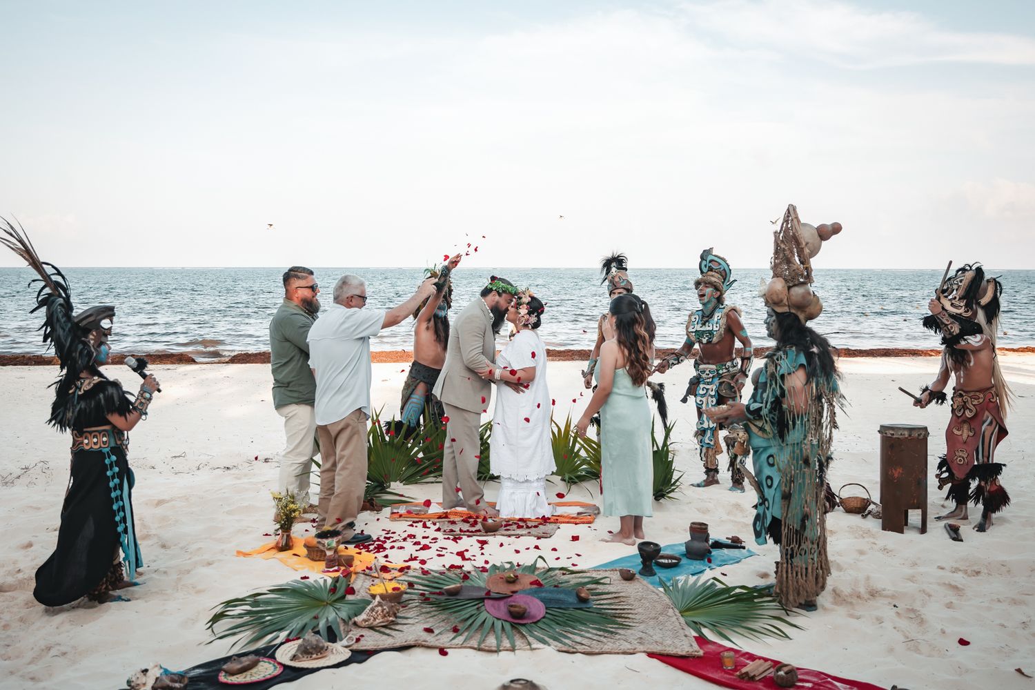 Couple exchanges vows in a beach wedding ceremony with Mayan performers in traditional costumes by the ocean.