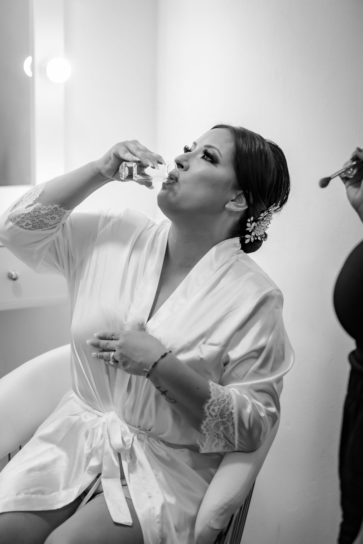 Bride in white lace robe drinking champagne while getting makeup done in black and white wedding preparation photo.