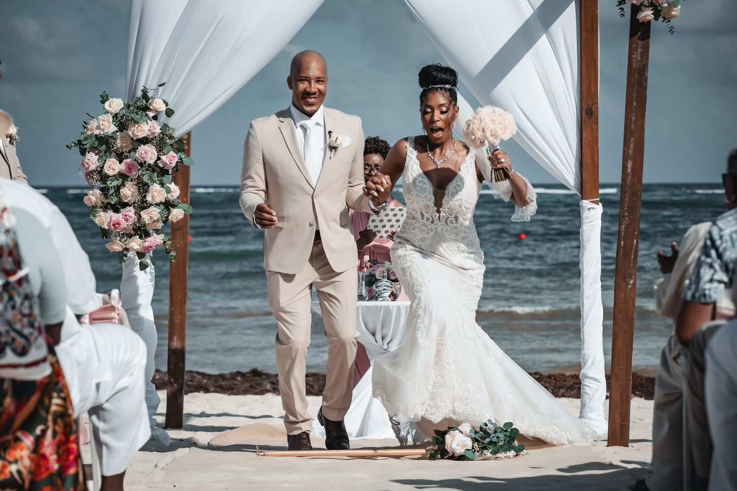 Bride and groom walking down beach wedding aisle with ocean backdrop and floral arch decorations.