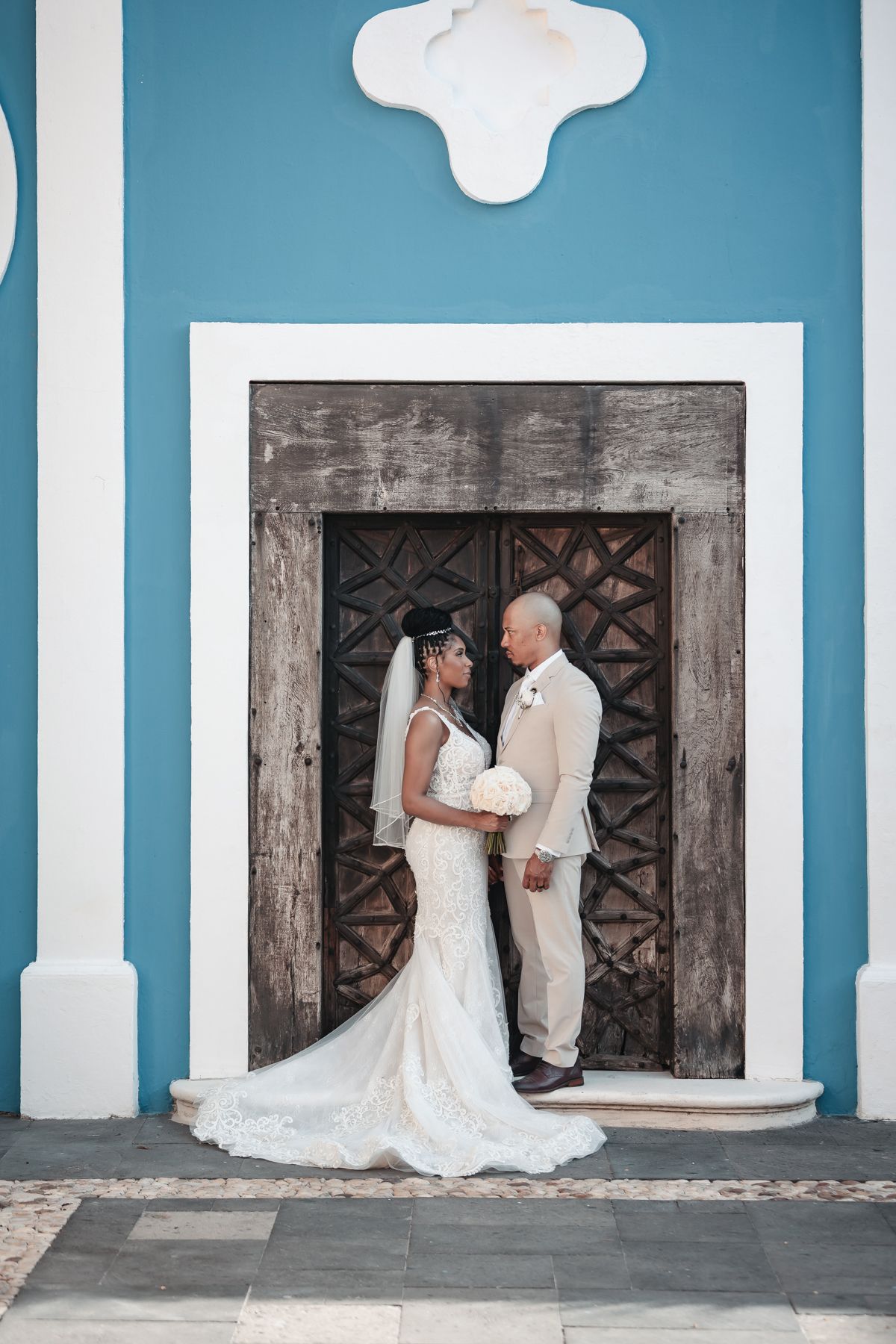 Bride and groom share a romantic moment in front of rustic wooden doors on a blue colonial building.