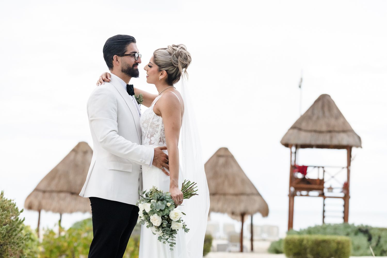 Bride and groom embrace at tropical beach wedding venue with thatched palapa huts in background.