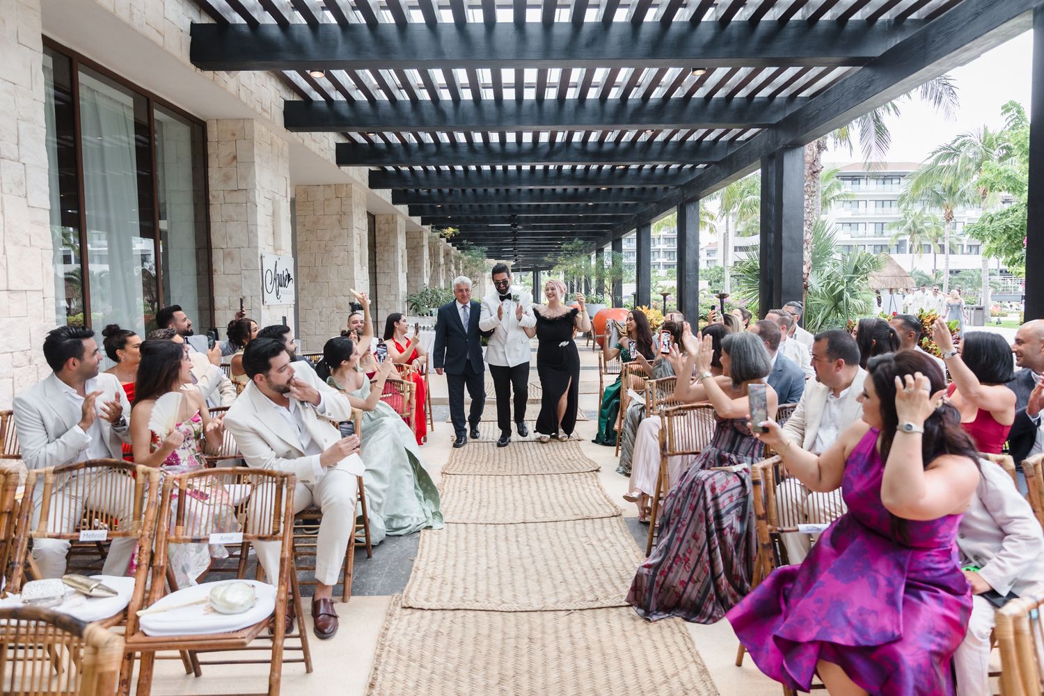 Wedding ceremony processional with guests seated outdoors under a pergola in tropical setting.