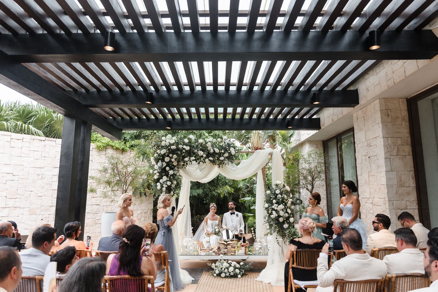 Wide-angle view of outdoor wedding ceremony under pergola with white floral arch and seated guests.