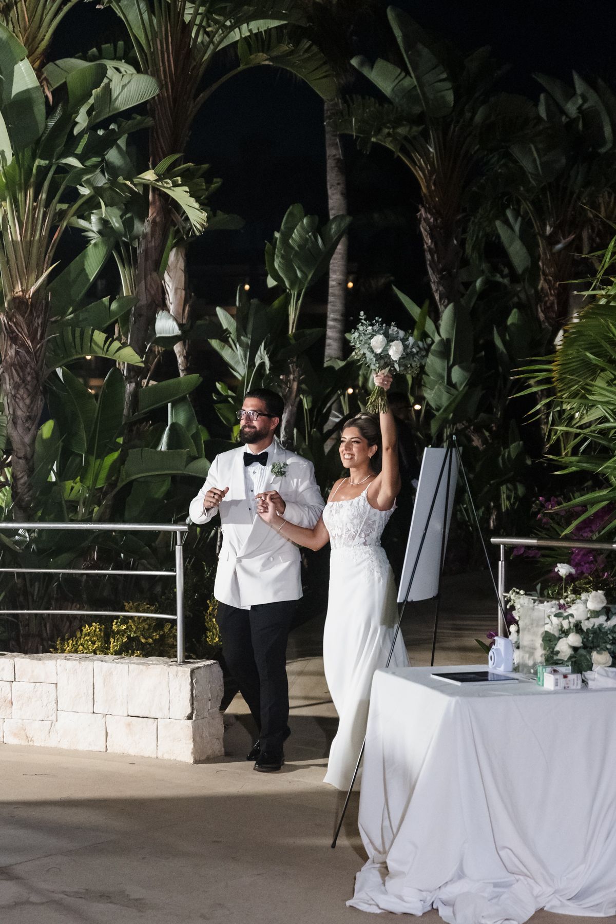 Bride and groom celebrate their wedding reception outdoors at night, surrounded by lush tropical greenery.