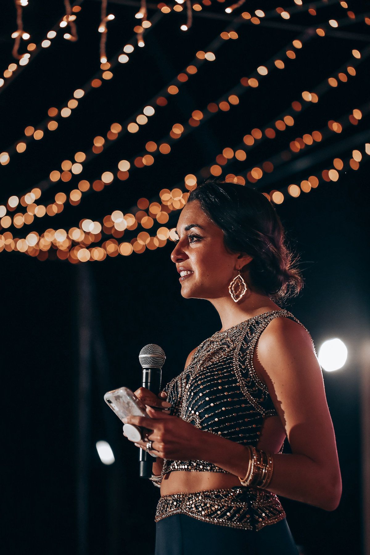 A speaker holds a microphone under decorative string lights at an evening event wearing a sparkly formal outfit.