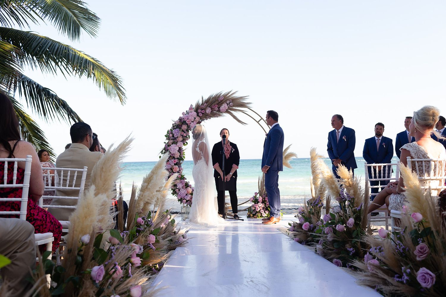 Wedding ceremony setup on a tropical beach with palm trees and white chairs overlooking the ocean.