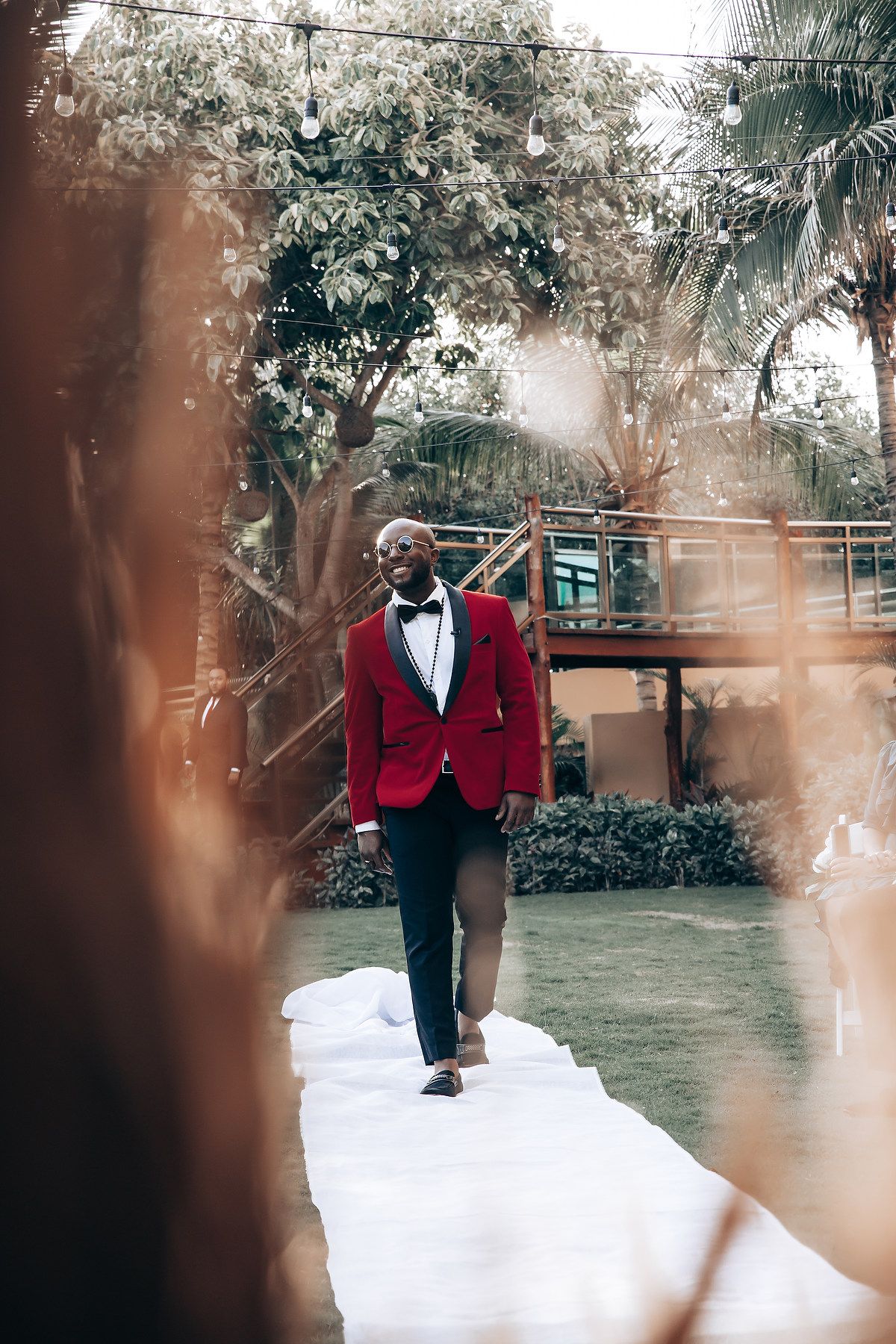 A person in a red velvet tuxedo jacket and black bow tie poses elegantly in a lush garden setting.