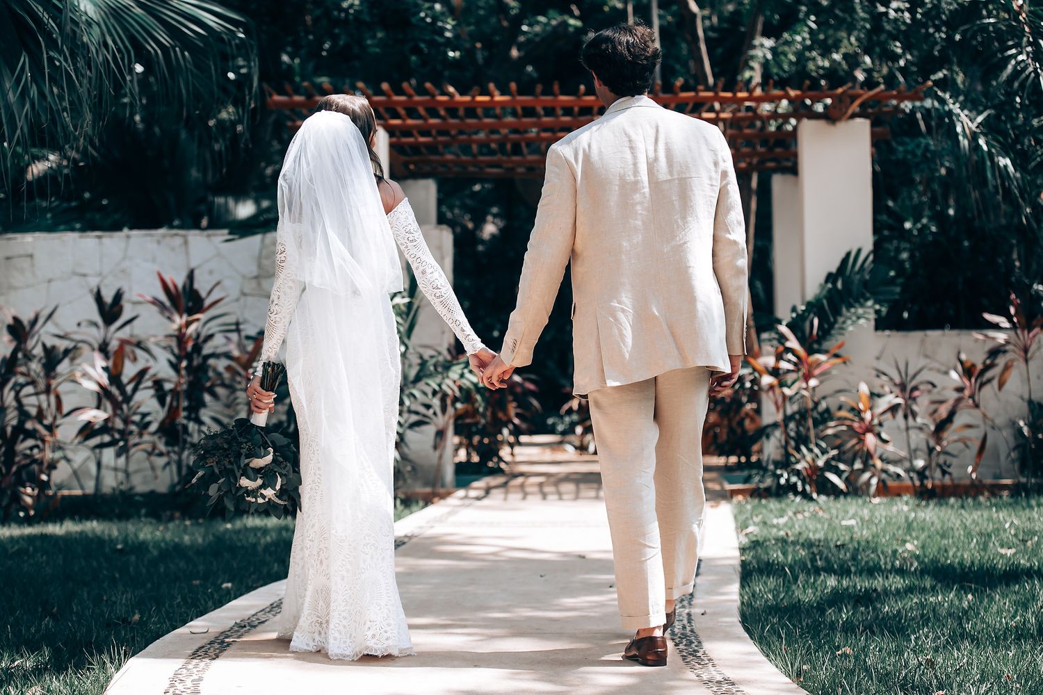 Couple walking hand in hand along a garden path in wedding attire.