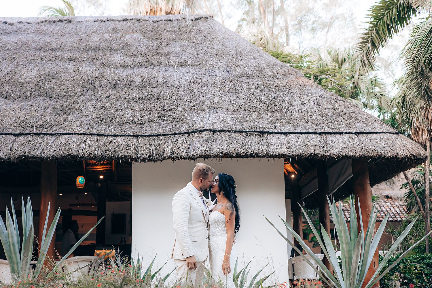 Wedding couple in front of a thatched-roof tropical bungalow.