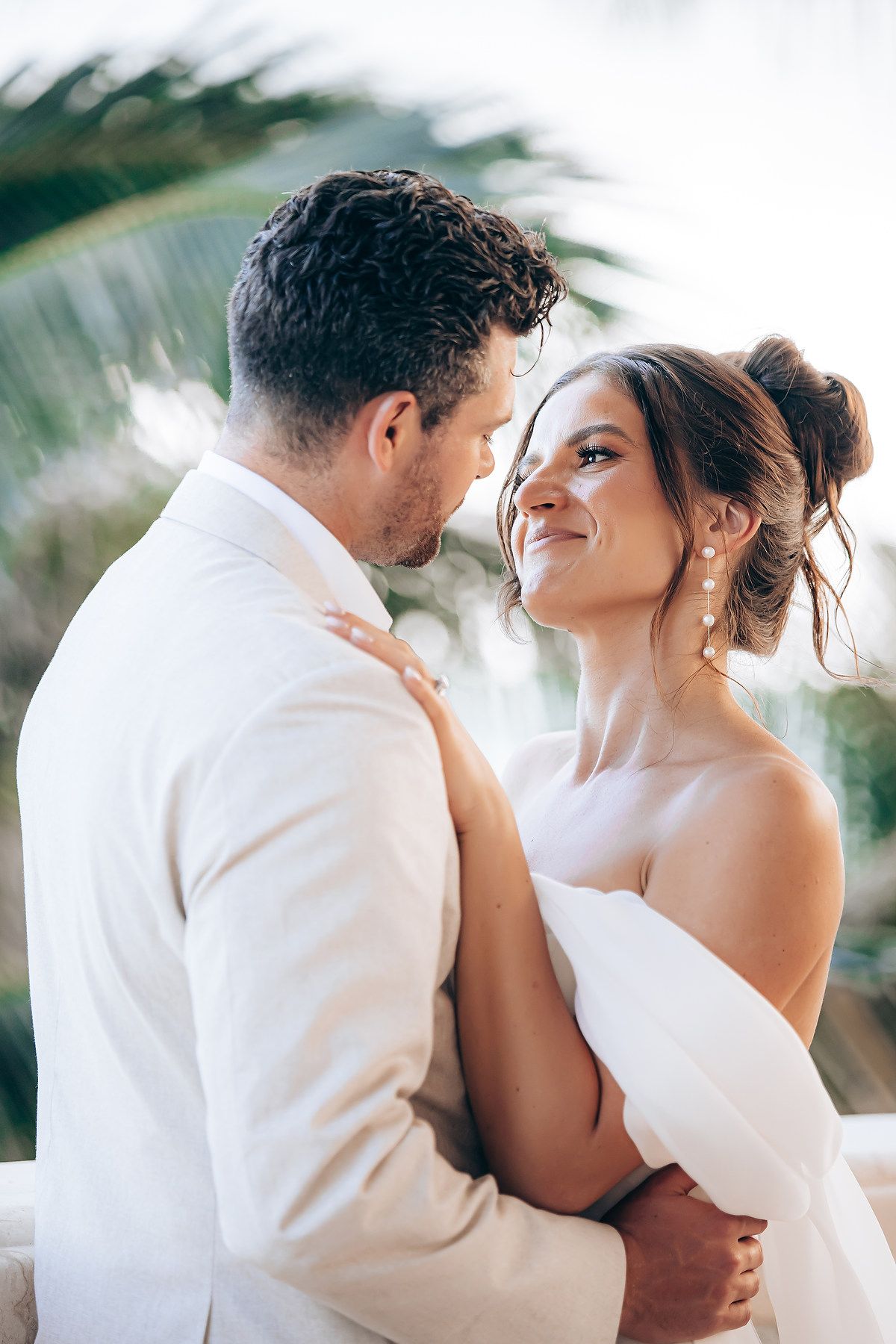 Romantic wedding couple embraces outdoors in tropical setting wearing white attire.
