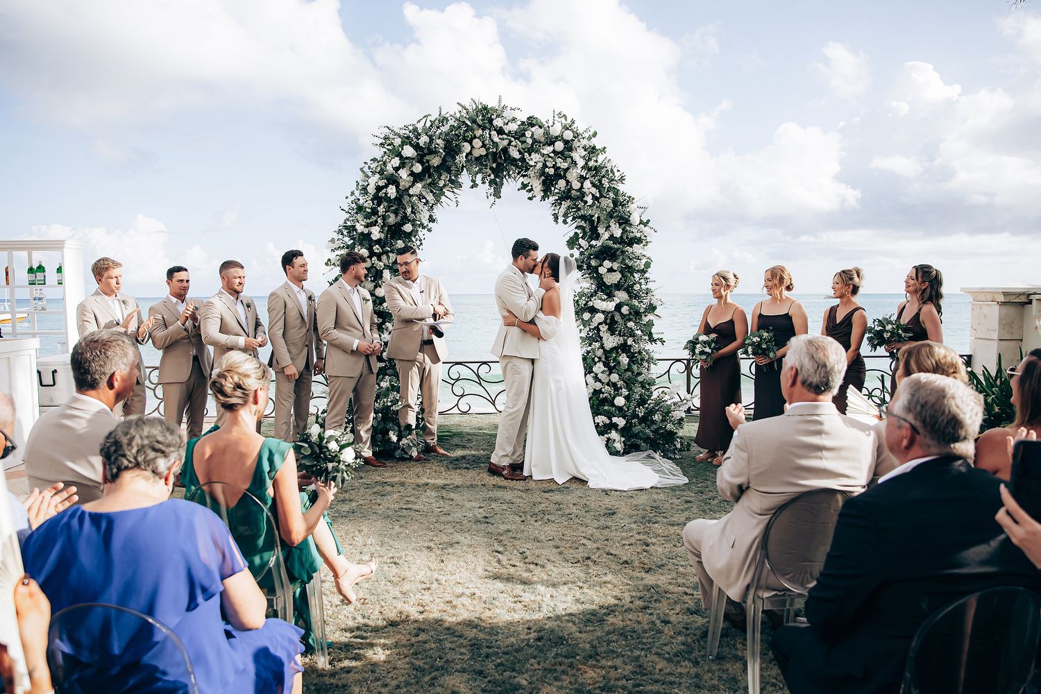 Wedding ceremony guests gather outdoors under palm trees and blue skies at a tropical beach venue.