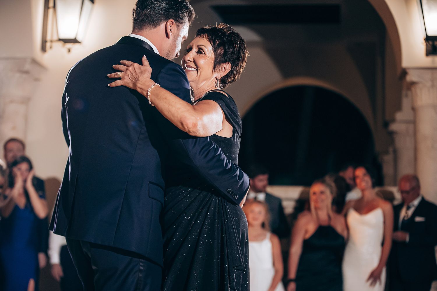 A couple shares their first dance at a wedding reception while guests watch and take photos in the background.