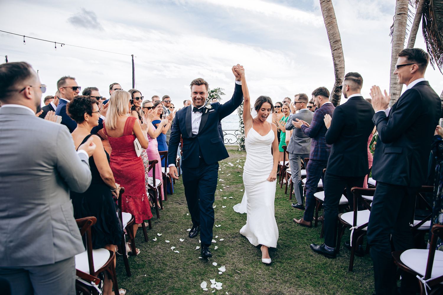 Newlyweds celebrate with raised arms while walking down outdoor aisle as guests cheer during oceanfront ceremony.