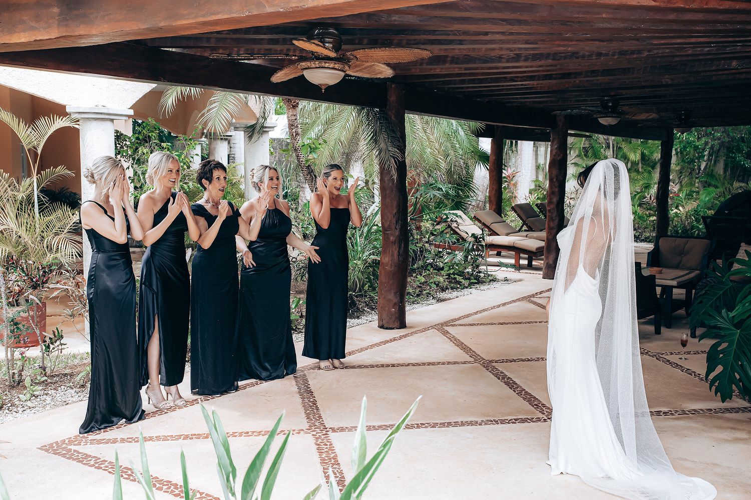 Wedding party members in formal attire gather under a covered outdoor walkway with tropical landscaping.
