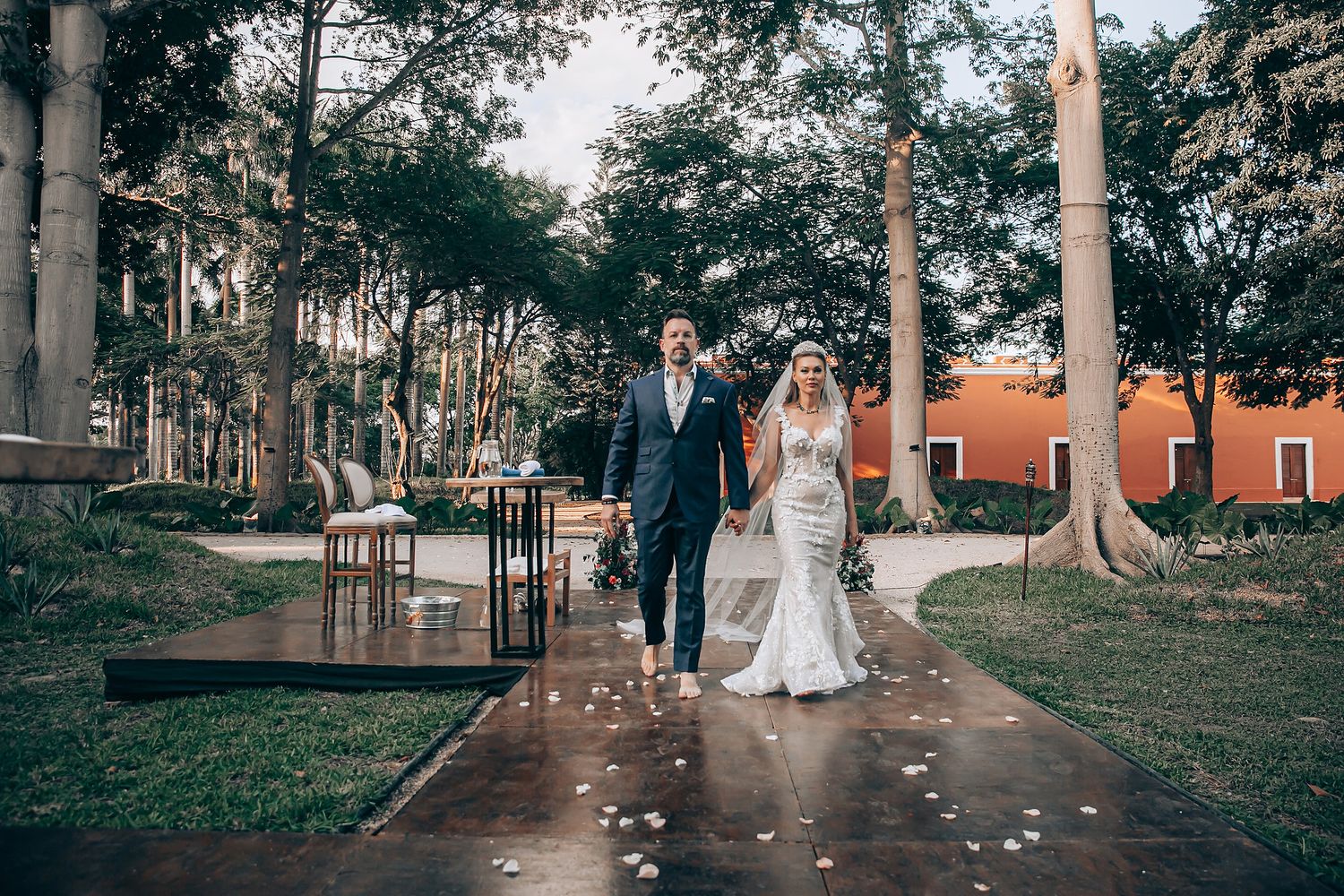 A wedding couple walks along a tree-lined path with scattered rose petals and vintage decor at an outdoor venue.