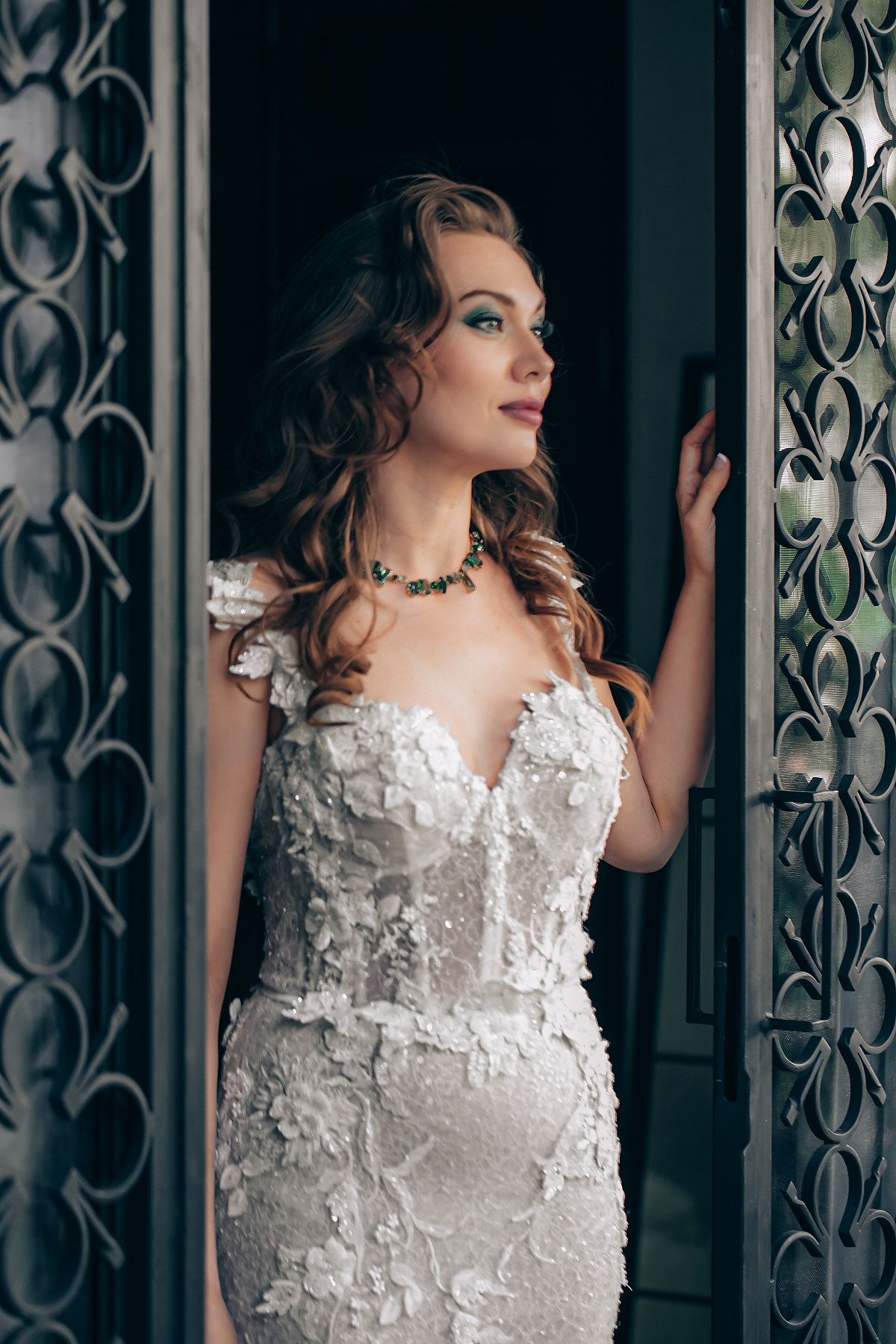 A bride in an elegant white lace wedding dress poses against an ornate door with decorative metalwork details.