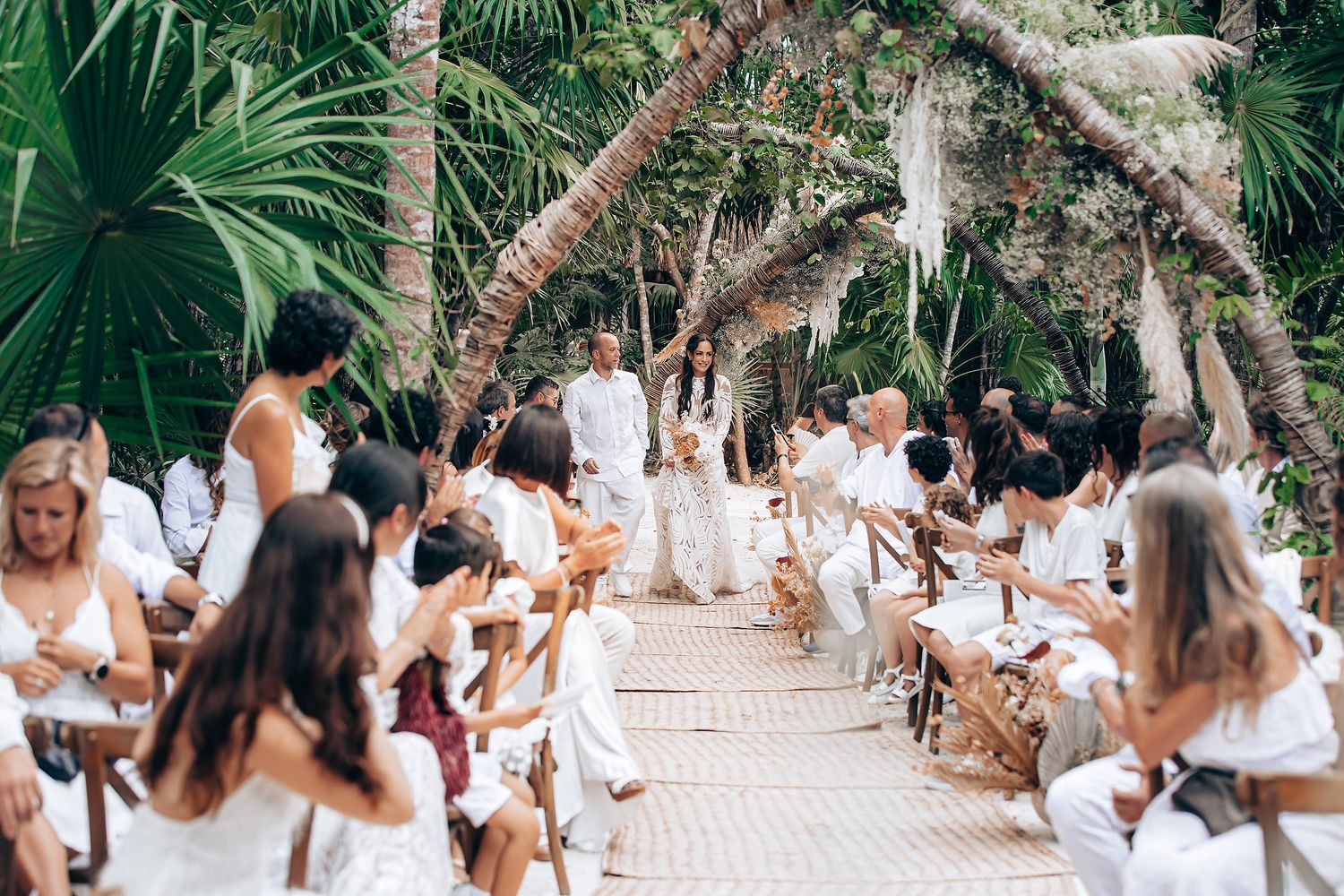 Outdoor wedding ceremony in a tropical garden setting with guests seated on wooden chairs along a stone pathway.