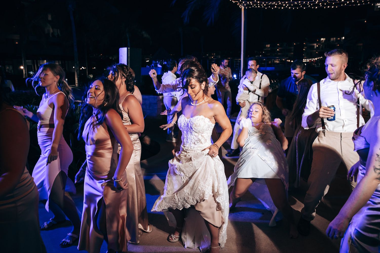 Wedding party dancing and celebrating under blue lighting at nighttime reception.