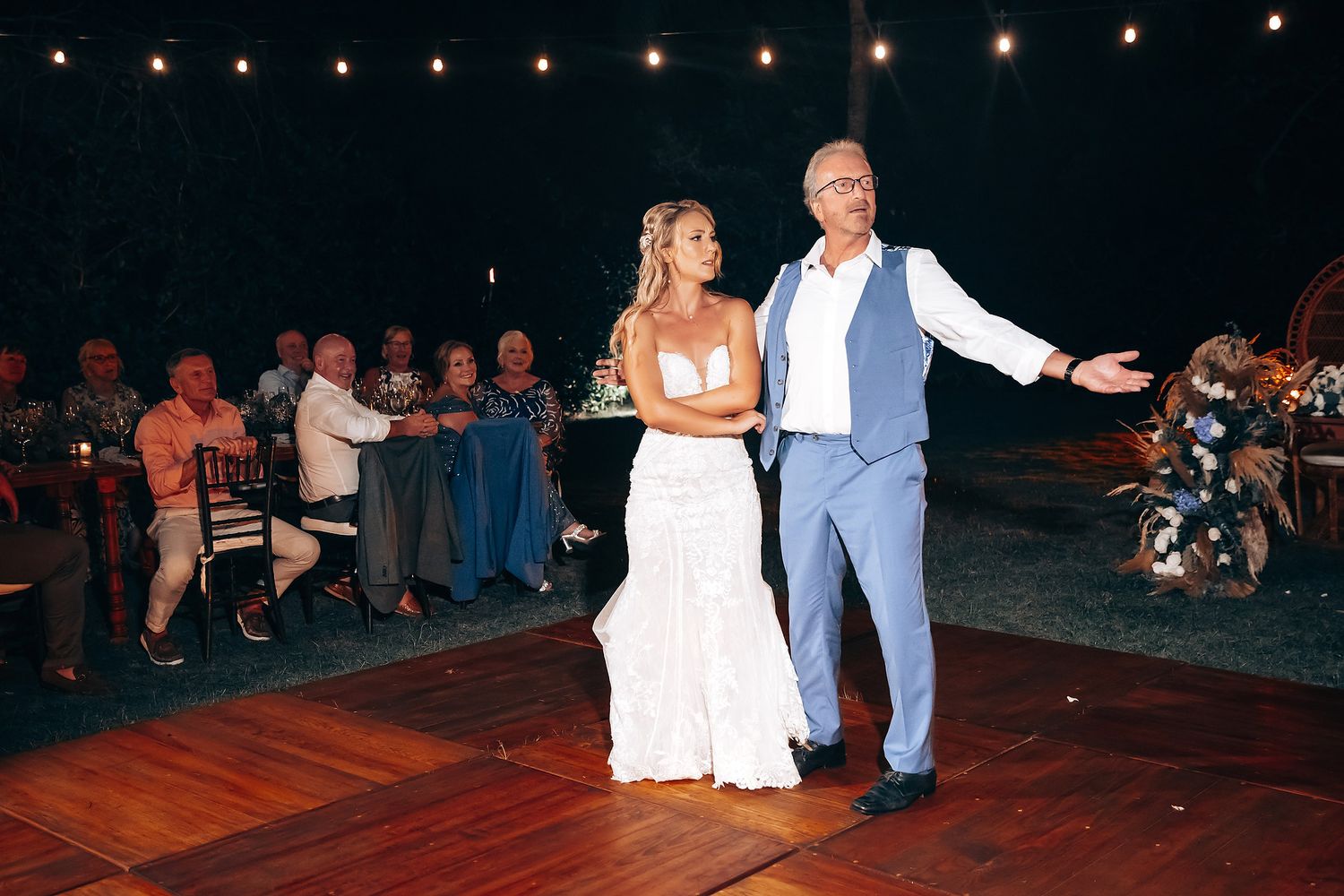 A couple performs their first dance on an outdoor wooden deck under string lights at night.