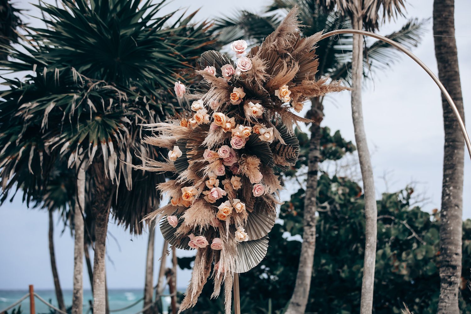 Tropical wedding ceremony setup with dried palm fronds and floral arrangements under palm trees.