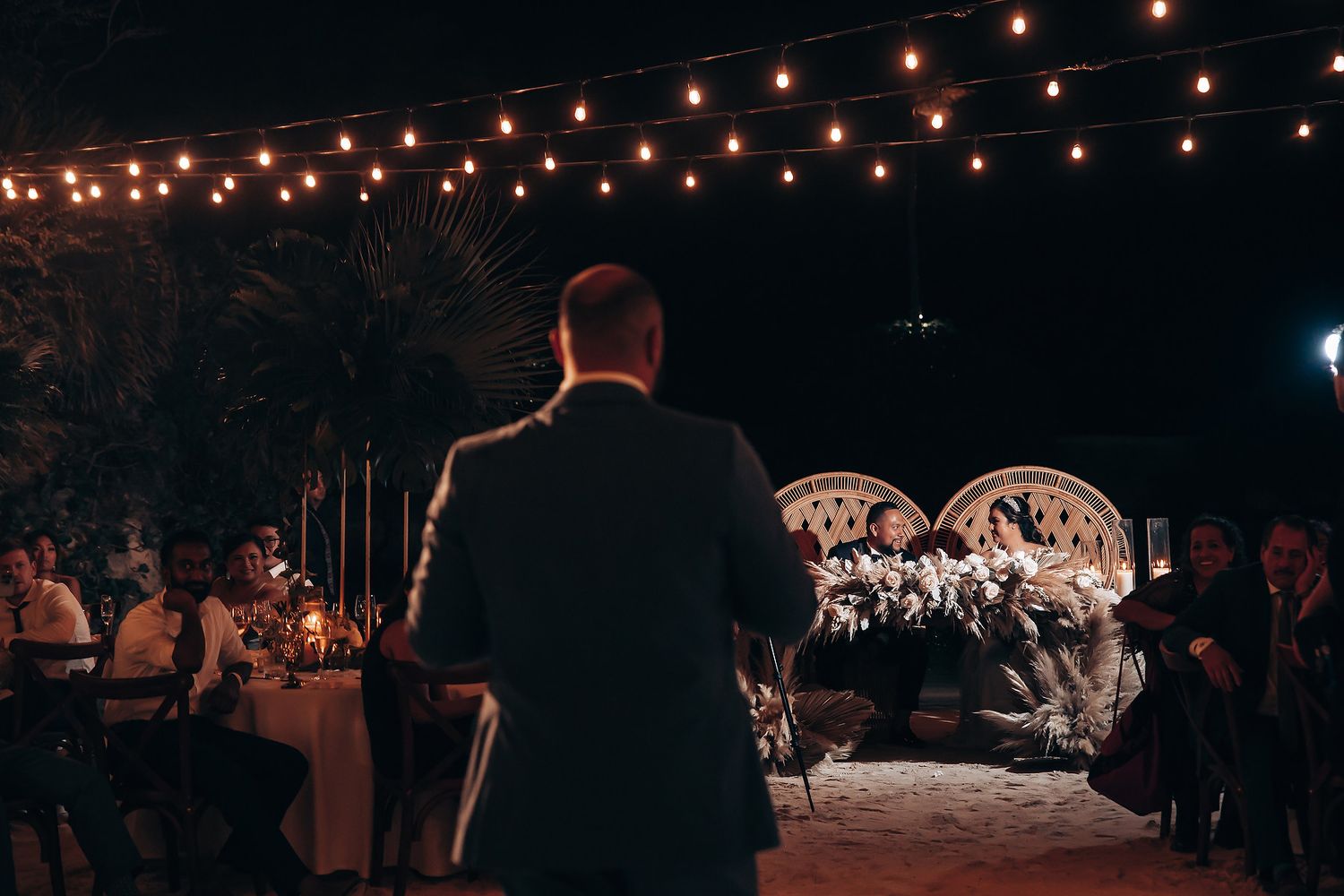 Warm string lights glow above a nighttime wedding reception creating an intimate atmosphere.