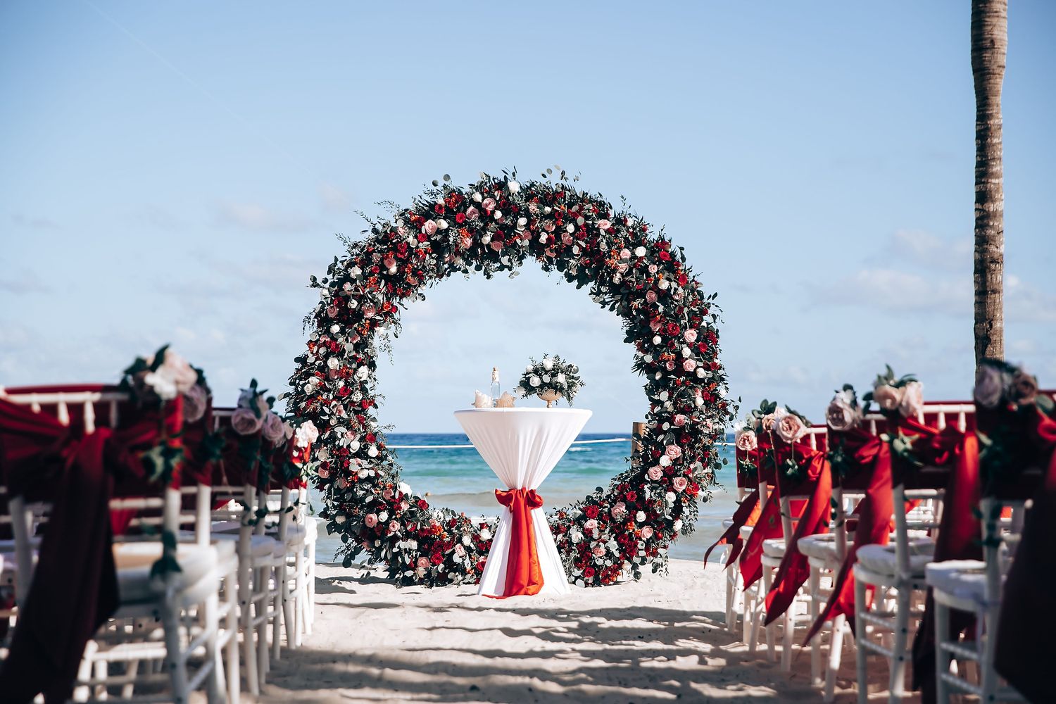 Tropical beach wedding ceremony setup with red floral arch and white chairs.