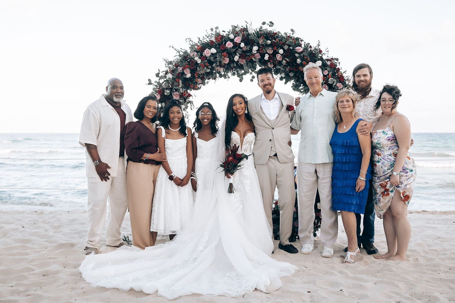 Wedding party in light colored attire poses together on a sunny beach with ocean waves in the background.