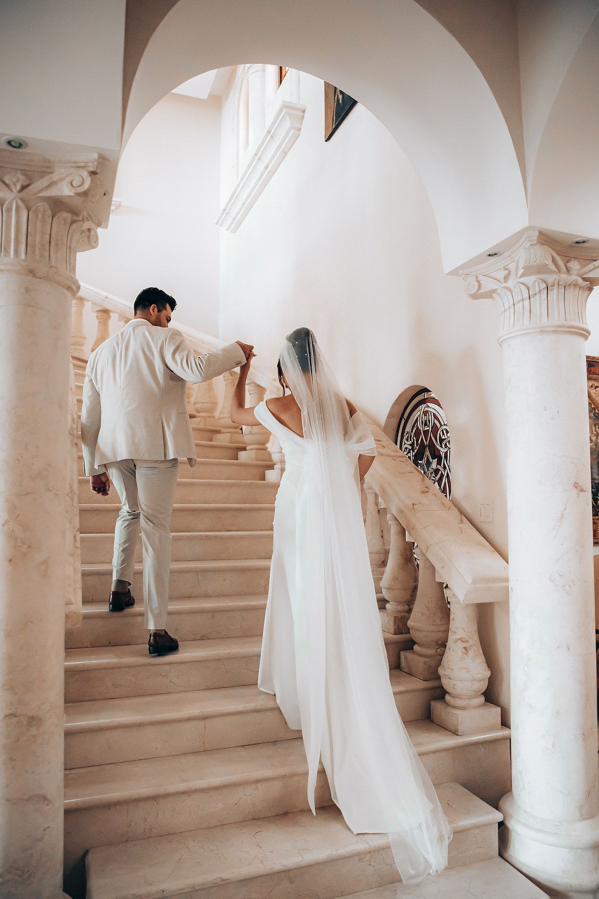 A couple ascends an elegant white marble staircase with classical columns and architectural details.