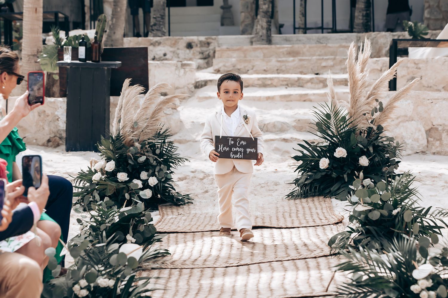 Young ring bearer walking down aisle at beach wedding holding wooden sign.