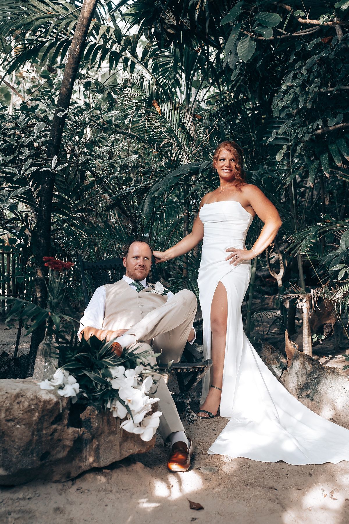 Couple takes casual wedding photos sitting on rocks in a tropical garden setting.