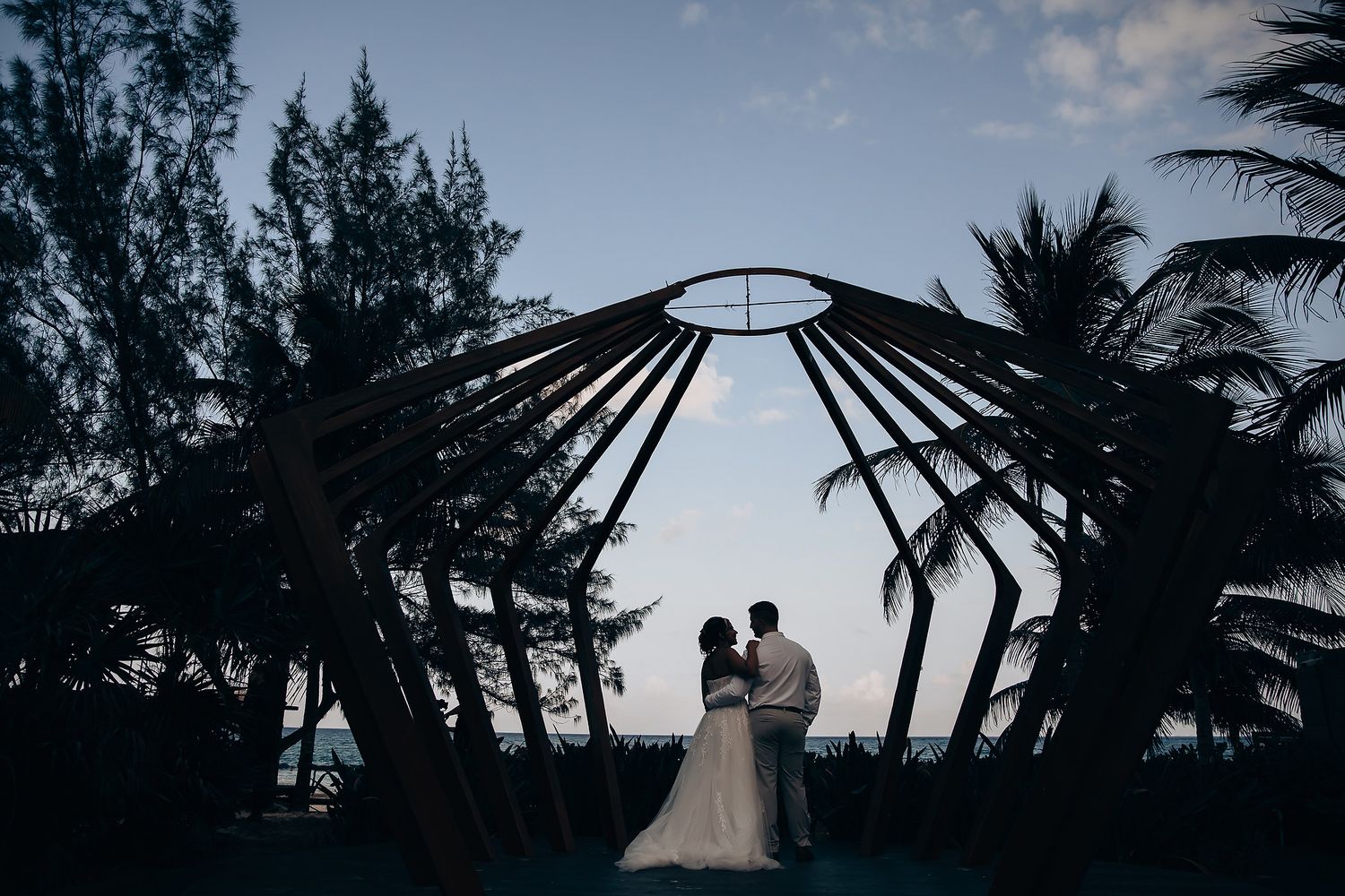 A silhouetted couple stands under a modern wooden pavilion at dusk surrounded by palm trees.