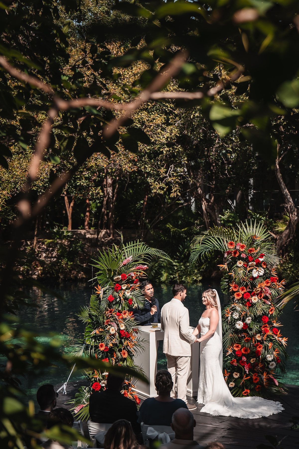 Intimate outdoor ceremony by turquoise water with dramatic red floral arch installations.