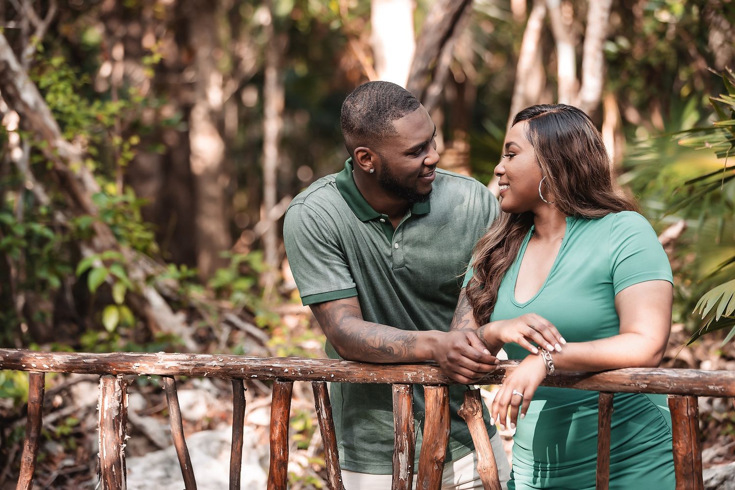 A couple shares intimate moments while leaning against a wooden railing in a forest setting.