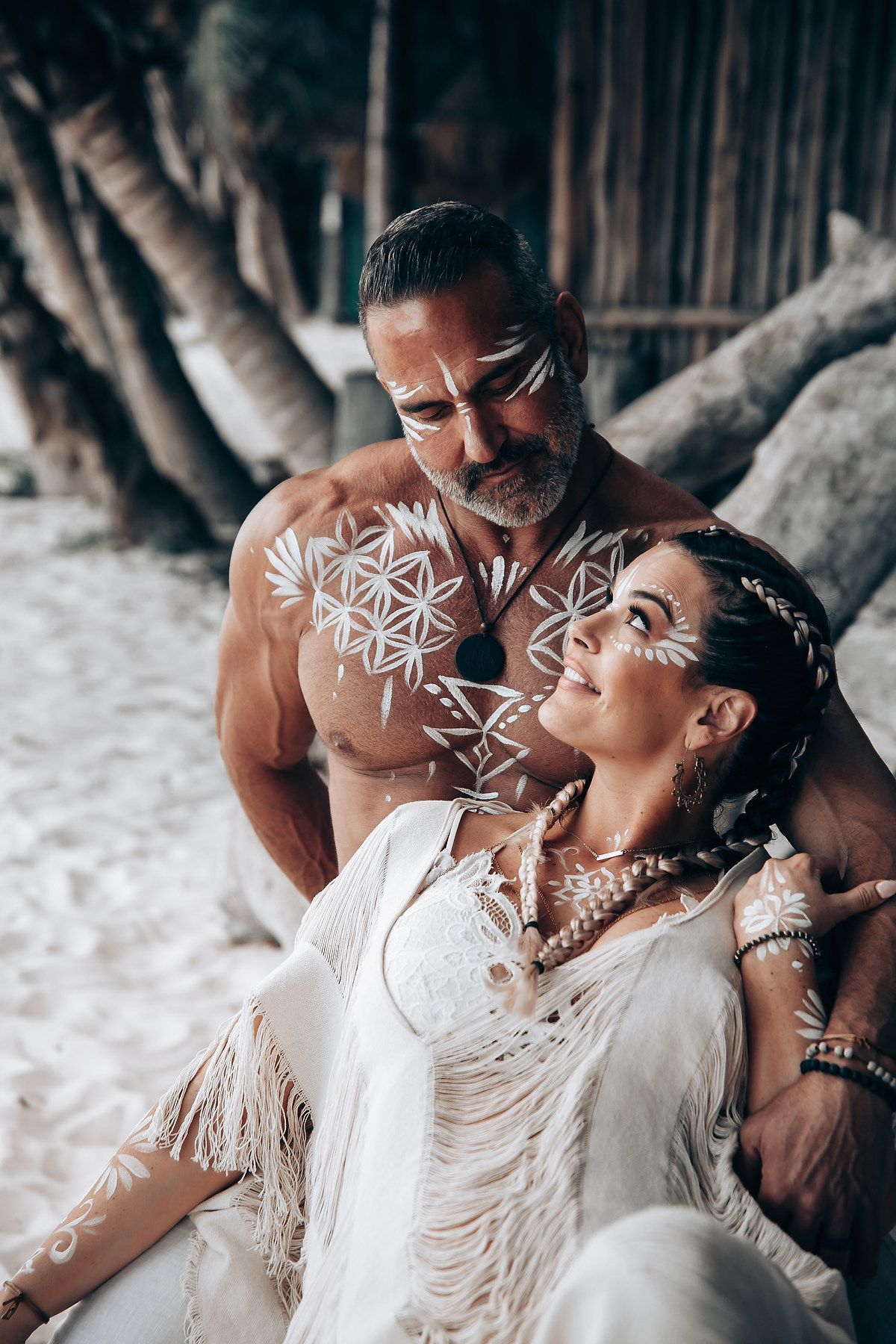 An intimate beach portrait with tribal body paint and flowing white fabric against palm trees.
