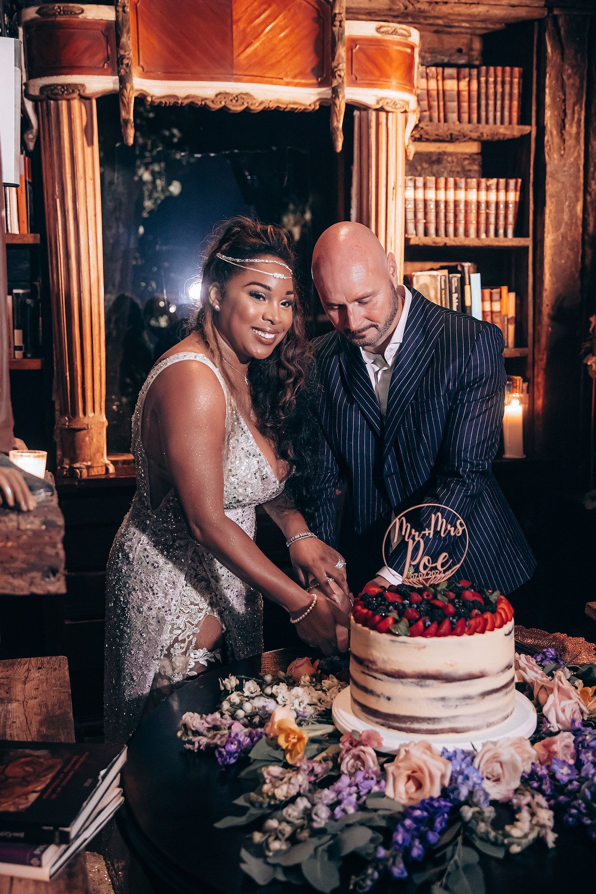 A couple cuts their elegant wedding cake adorned with berries and floral decorations in a vintage library setting.