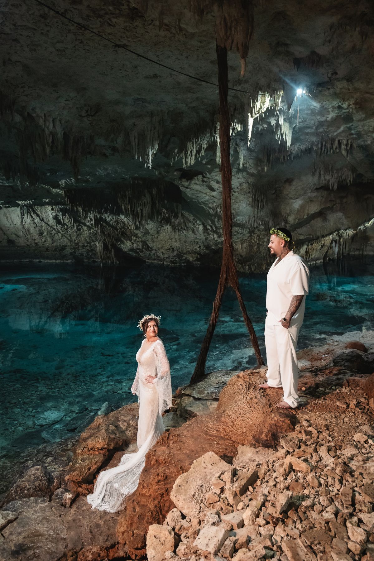 Bride in flowing white dress poses gracefully along rocky cave ledge with glowing blue water below.