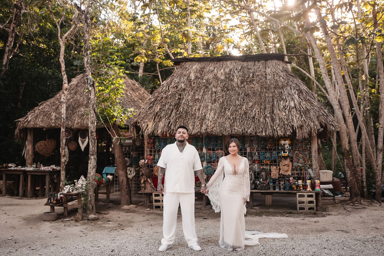 Wedding couple poses in front of rustic thatched-roof buildings and lush tropical trees during sunset ceremony.