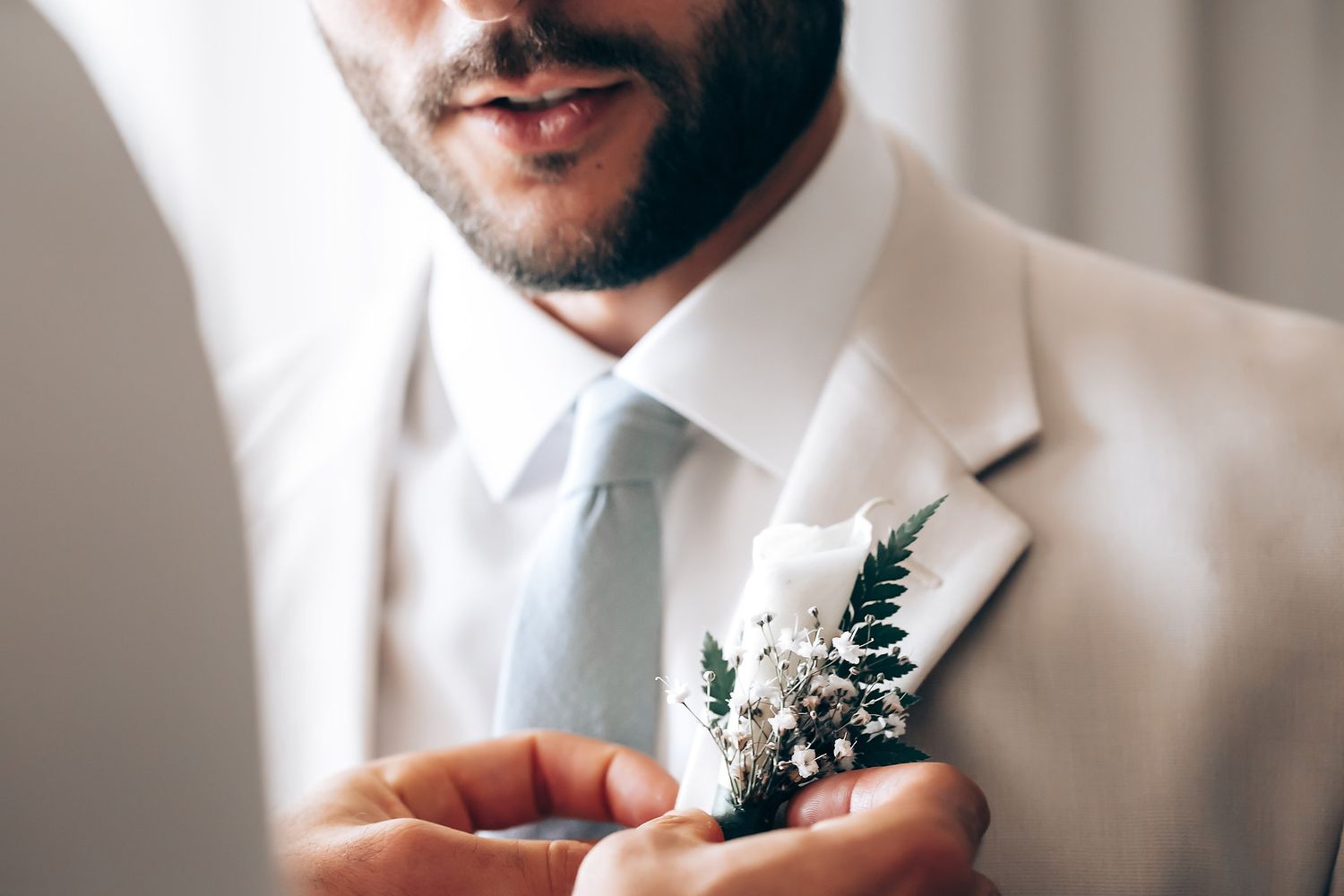 A delicate boutonniere with ferns and white flowers is pinned to a white suit jacket.