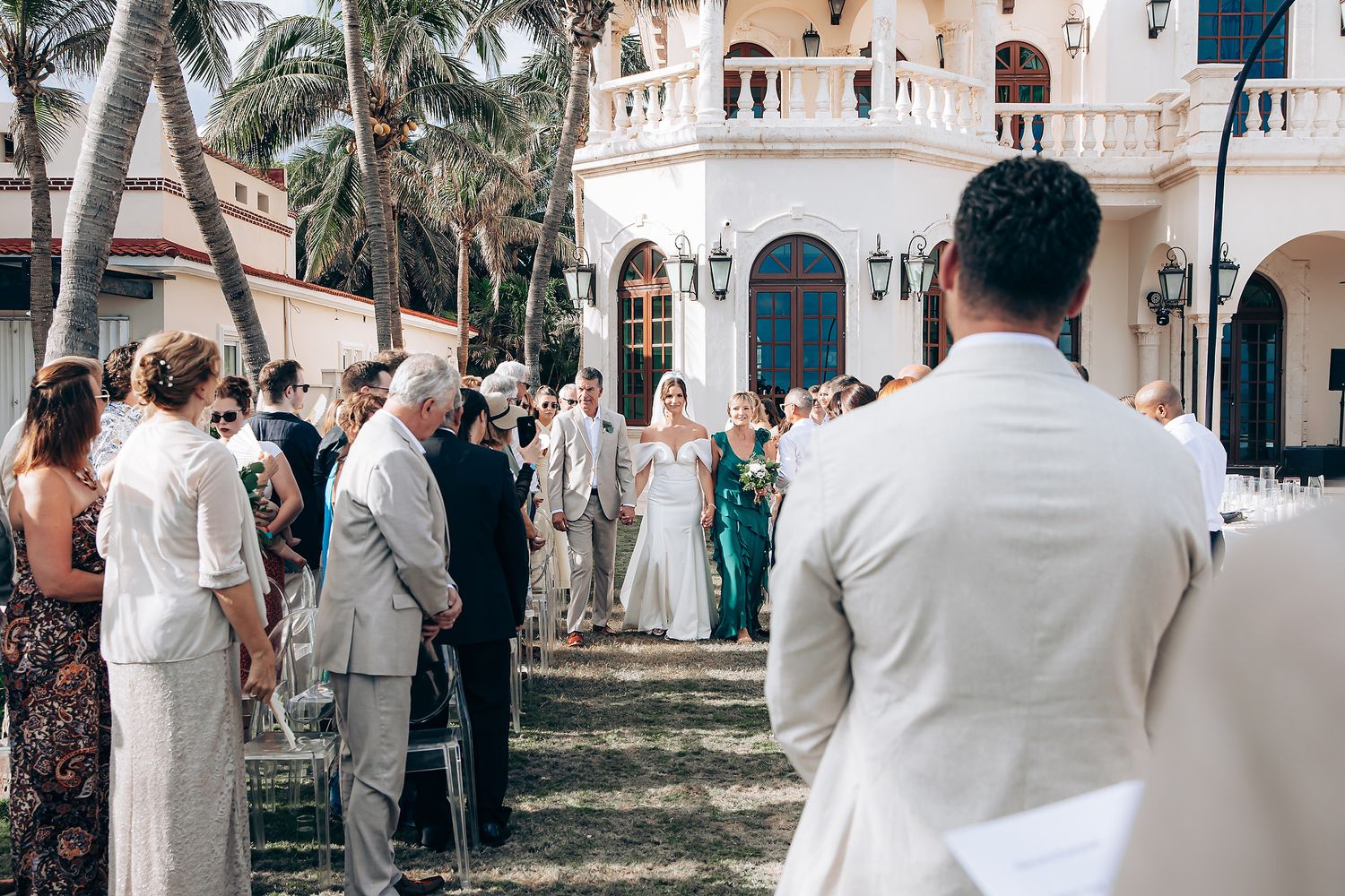 Wedding guests in white and formal attire gather outside a Mediterranean-style venue.