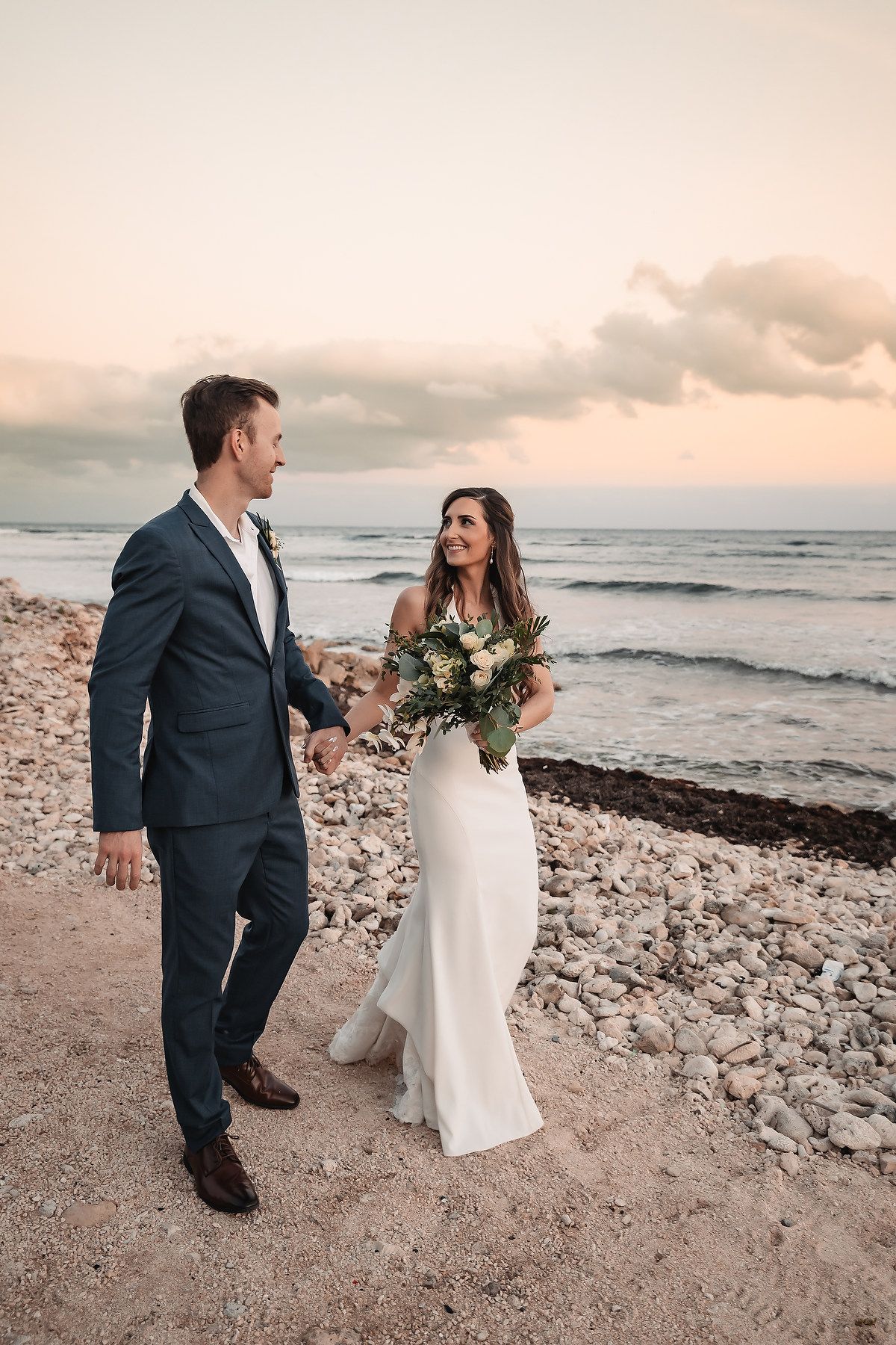 Couple walking along rocky beach shoreline at sunset during wedding photoshoot.