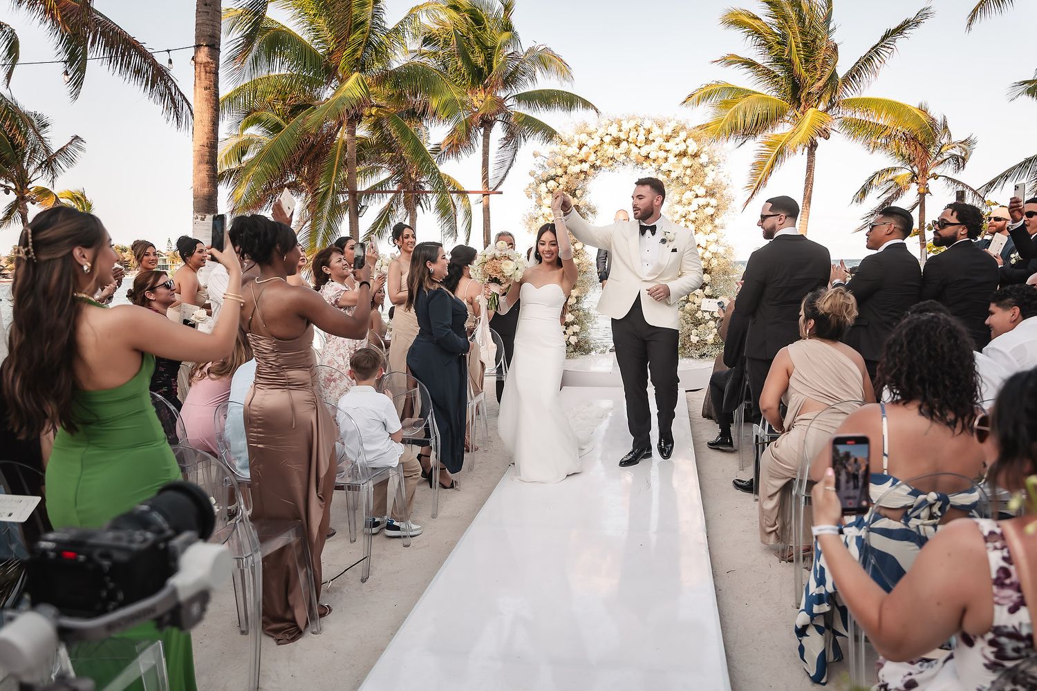 Wedding ceremony on a tropical beach with palm trees and white decor featuring a circular floral arch.