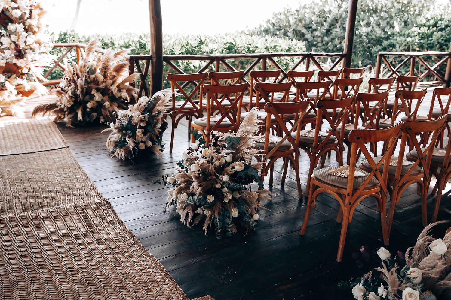 Beachside wedding ceremony setup with wooden crossback chairs and tropical floral arrangements.