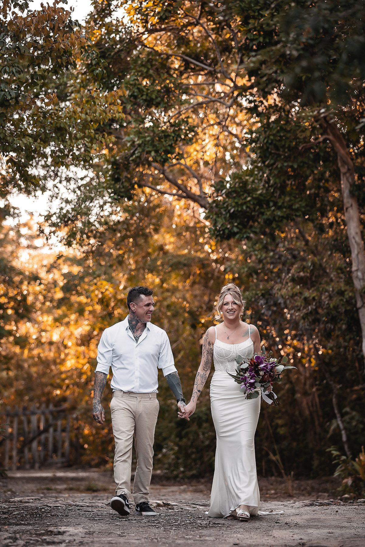 Smiling newlyweds walk hand in hand through golden-lit forest, groom tattooed in white shirt, bride in fitted gown.