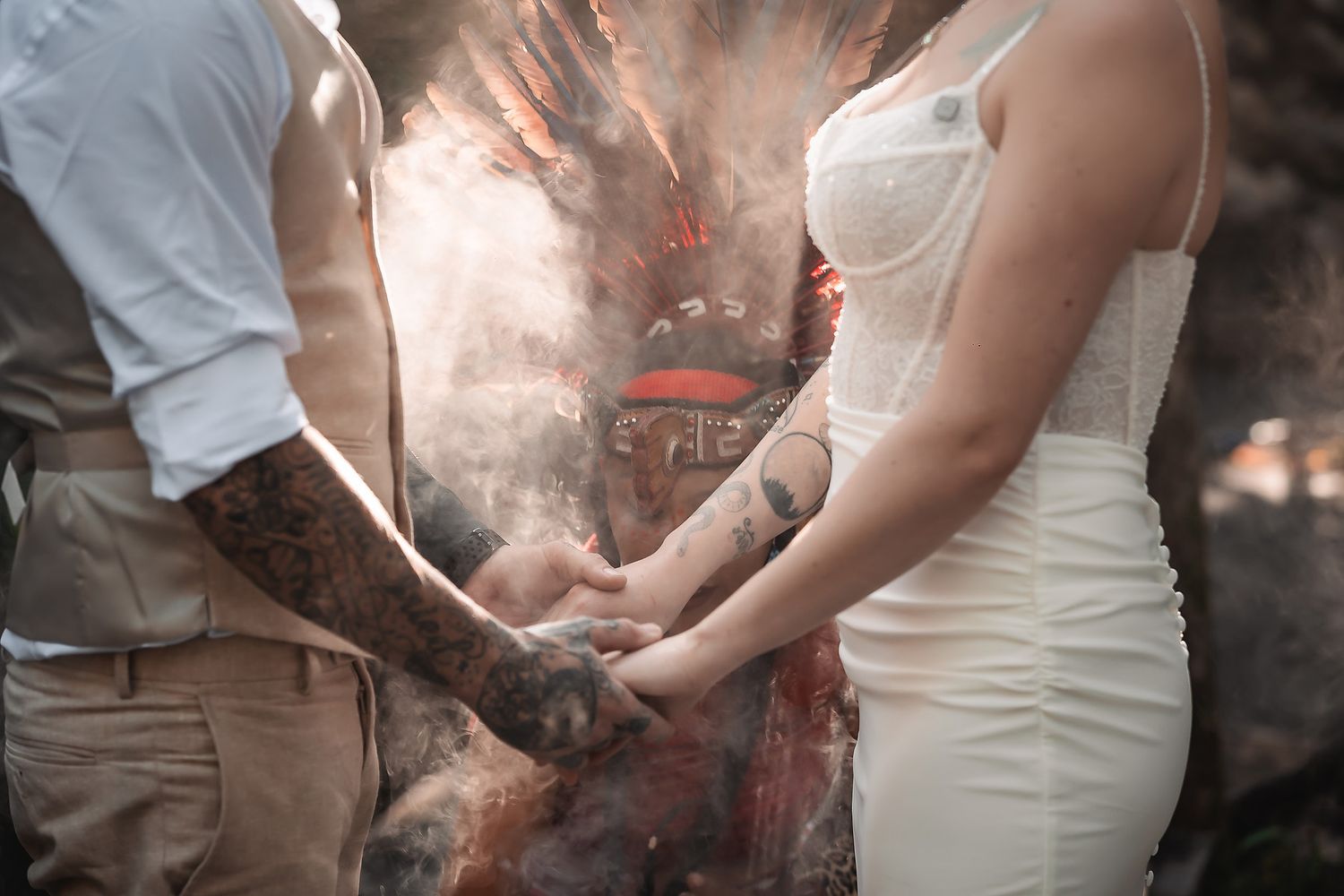 Tattooed couple holding hands during an outdoor wedding ceremony with smoke and rustic backdrop.