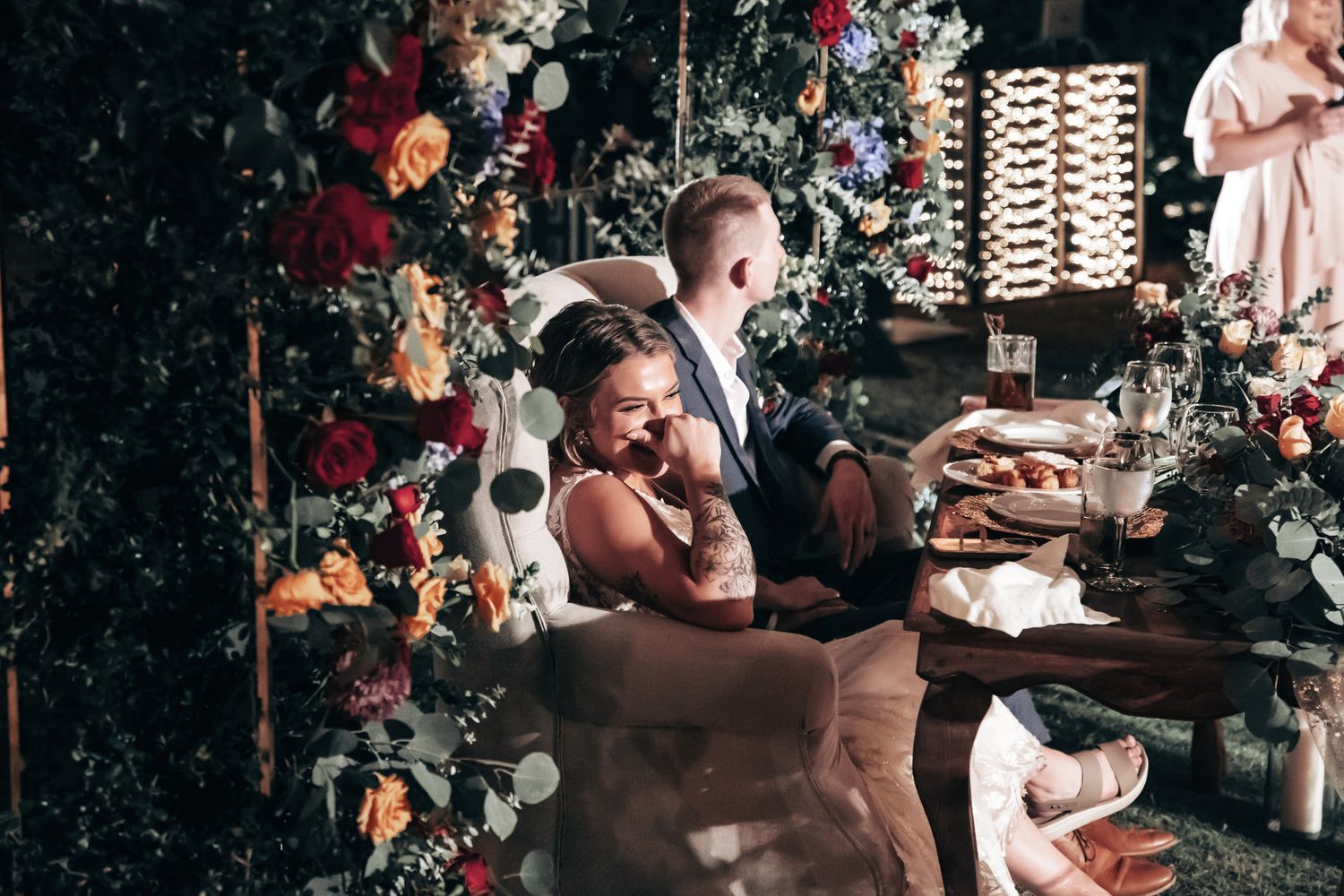Bride and groom laugh together at their floral-adorned sweetheart table during evening wedding reception.