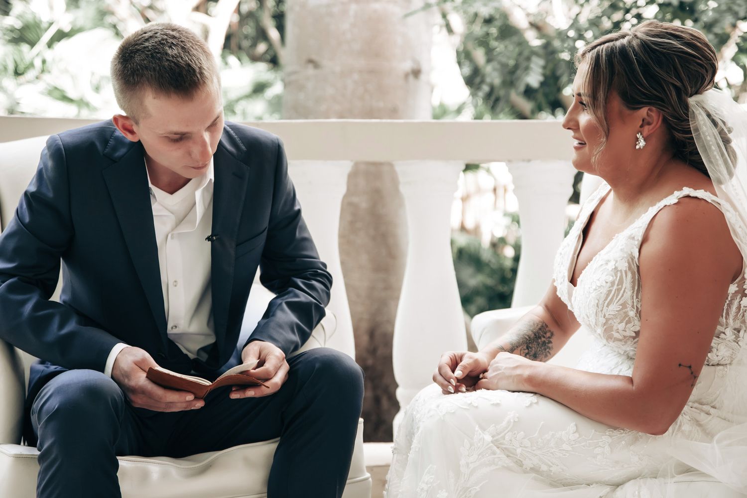 Groom reading heartfelt wedding vows from a card while seated next to his bride during an intimate outdoor ceremony.