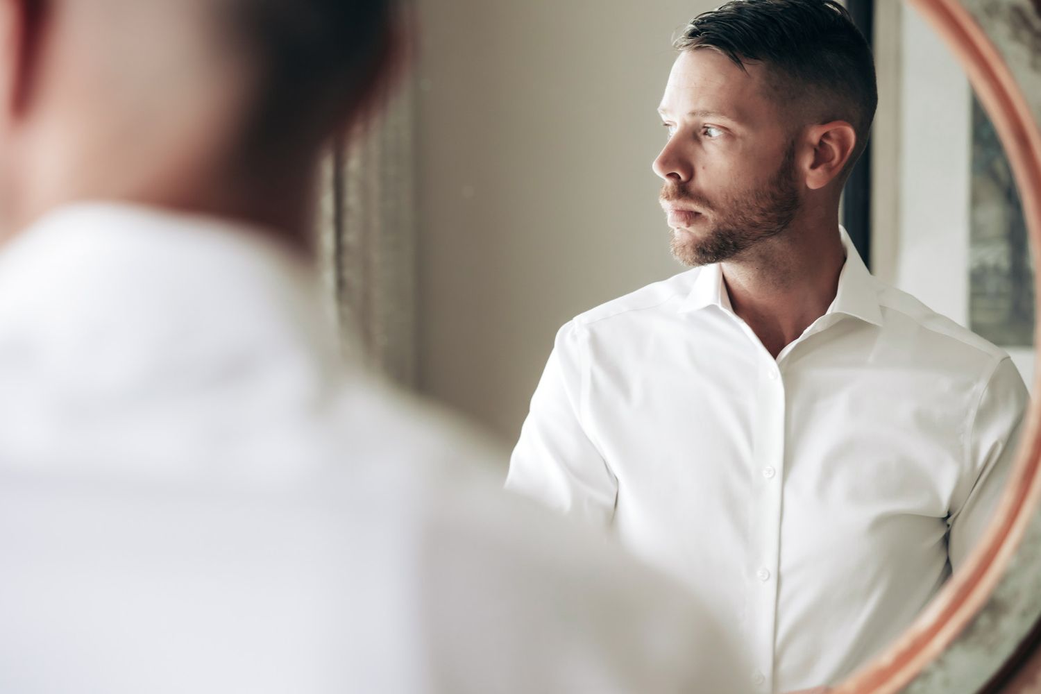 A well-groomed man in a white dress shirt gazes thoughtfully into a round mirror while getting ready.